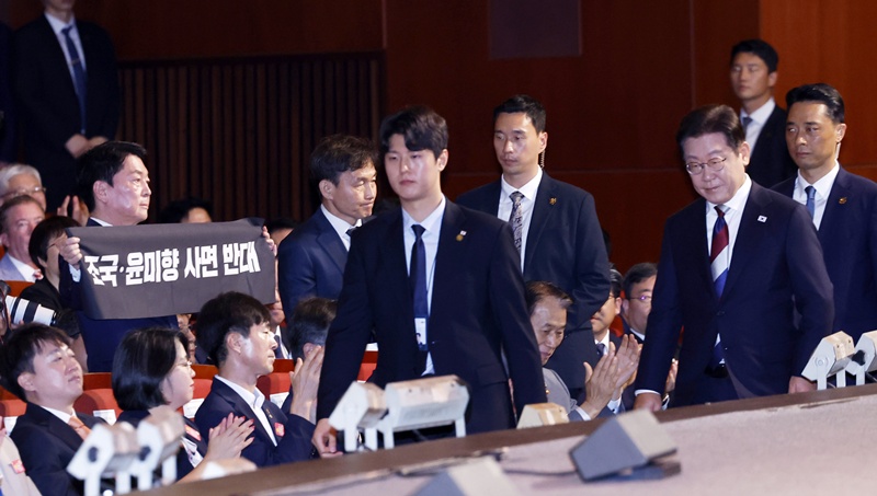 이재명 대통령이 지난해 8월 15일 서울 종로구 세종문화회관에서 열린 제80주년 광복절 경축식에서 경축사를 마치고 이동하고 있는 가운데 안철수 국민의힘 의원이 조국, 윤미향 사면 반대 피켓을 들고 있는 모습이다. / 뉴시스